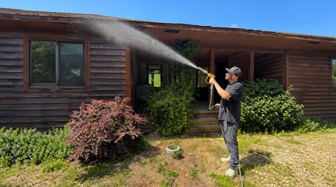 Person spraying water with a hose on a wooden house roof, with green shrubs and overgrown lawn visible.