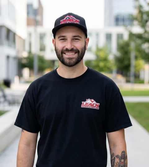 Smiling man wearing black cap and t-shirt with logo, standing on outdoor pathway with buildings in background